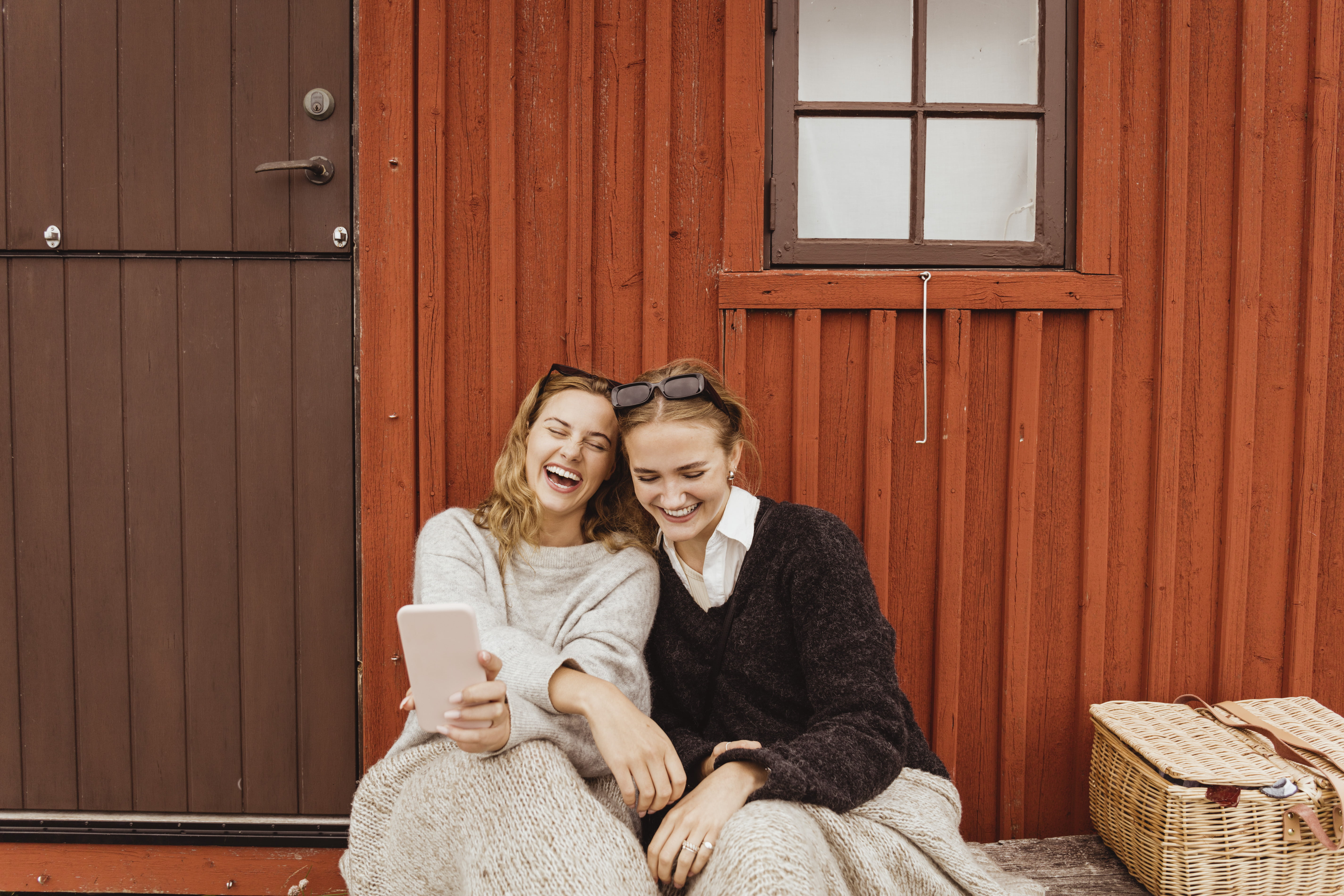 To damer ser på mobilen mens de sitter foran en rød husvegg